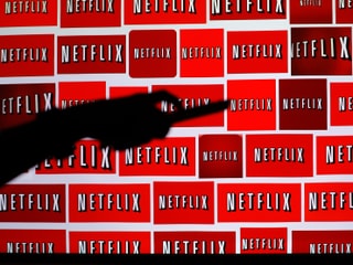 Netflix auf diversen Monitoren, davor eine Hand mit einer Fernbedienung