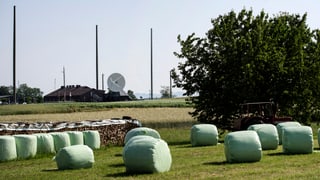 Ein Feld mit in plastik eingepackten Strohballen, im Hintergrund ist eine grosse Satellitenantenne zu erkennen.