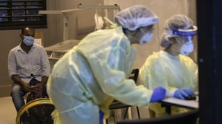 Zwei freiwillige Pflegehelfer in Schutzanzügen testen einen Mann auf das Coronavirus, sie schauen auf einen Bildschirm.