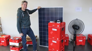 Mann hält Solarpanel, rote Kisten daneben.
