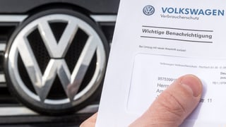Ein Mann hält einen Brief von VW in der Hand, im Hintergrund ein Auto mit dem VW-Logo.