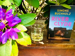 Der Krimi «River Clyde» von Simone Buchholz steht zusammen mit einem Glas Whiskey unter einem Rhododendronbuschunter