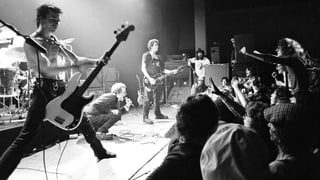 Eine Punk-Band steht auf der Bühne, ein Mann im Publikum macht ein Foto. Eine Punk-Band steht auf der Bühne, ein Mann im Publikum macht ein Foto.