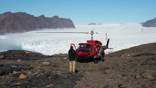 Ein roter Bell-Helikopter auf felsigem Grund. Im Hintergrund die Eisfläche des Bowdoin-Gletschers.