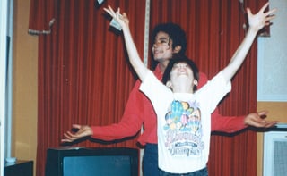 Michael Jackson steht hinter einem Jungen, der Geldscheine zu werfen oder zu fangen scheint. Die beiden lachen.