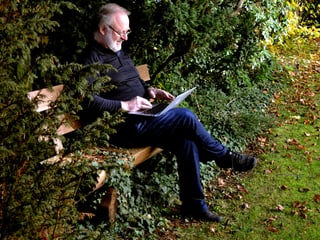 Ein Mann in dunkler Kleidung sitzt mit einem Laptop auf den Knien auf einer Holzbank, leicht versteckt hinter ein paar Tannenbaumzweigen.