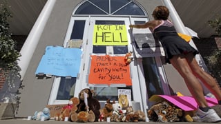 Plakate vor der Arztpraxis der Jägers. Auf einem steht auf Englisch geschrieben: «Schmore in der Hölle».