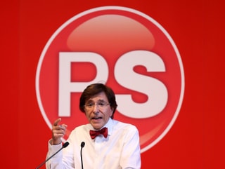 Elio di Rupo mit Fliege vor einer roten Wand mit dem Logo der Parti Socialiste.