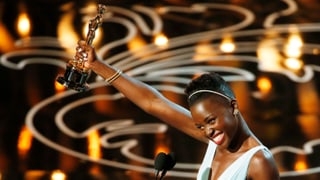 Lupita Nyong'o strahlt und hält den Oscar in die Luft.