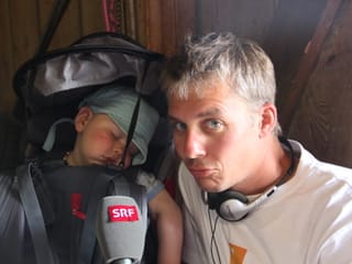 Reto Scherrer kniet neben einem Kleinkind, dass im Rucksack ein Schläfchen macht.