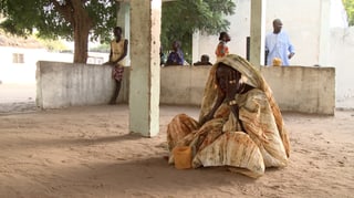 Eine Frau in afrikanischer Kleidung sitzt mit Händen vor dem Gesicht auf sandigem Boden.