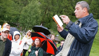 Andrzej Stasiuk liest aus einem Buch auf einer grünen Wiese, im Hintergrund hören Menschen zu mit teilweise aufgespannten Regenschirmen.