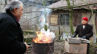 Zwei ältere Männer stehen neben einer brennenden Tonne in einem Vorstadt-Garten.