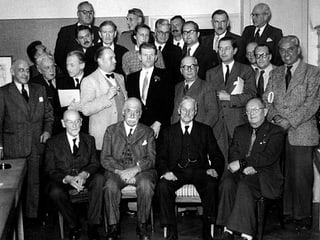 Eine Gruppe von Männern versammelt sich zu einem Gruppenfoto, in ihrer Mitte ein deutlich jüngerer Mann. Eine Gruppe von Männern versammelt sich zu einem Gruppenfoto, in ihrer Mitte ein deutlich jüngerer Mann.