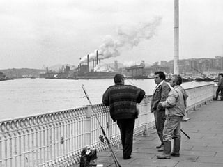 Fischer auf einer Brücke, dahinter Industriegebäude und rauchende Kamine