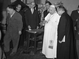 Der Papst in weisser Kutte auf einem schwarz-weiss-Bild, besucht 1959 die Mitarbeiter der Vatikanzeitschrift. Er ist umgeben von Mitarbeiternm, die ihn willkommen heissen. 