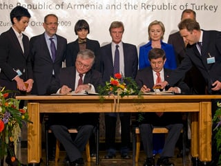 Frauen und Männer stehen hinter einem Tisch, an dem zwei Männer ein Dokument unterzeichnen.