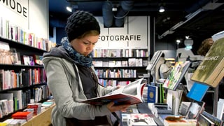 Eine junge Frau mit Mütze liest in einem Buchladen stehend ein Buch.