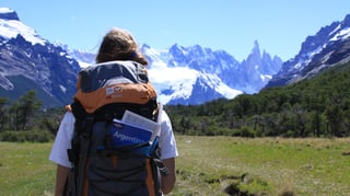 Eine Frau vor Schneebergen. In ihrem Rucksack steckt ein Reiseführer mit der Aufschrift "Argentina". 
