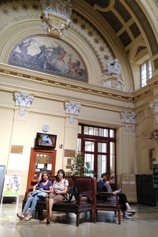 In einer geschmückten, hohen Eingangshalle sitzen ein paar Menschen auf Bänken.
