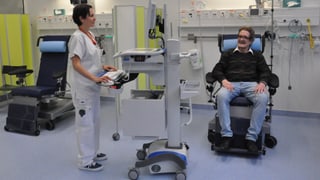 Ein Mann sitzt in einem Behandlungssessel und eine Pflegende steht an einem Computerwagen.