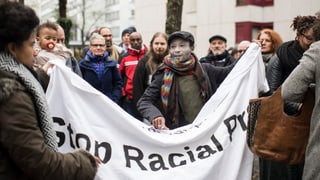 Eine Gruppe von Menschen hält ein Banner in die Kamera, darauf steht schwarz auf weiss: Stop racial profiling.