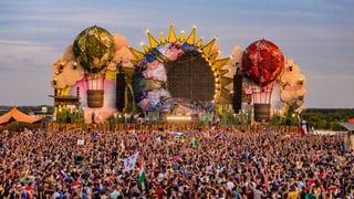 Viele Festivalbesucher vor einer grossen Bühne. Bühne ist eine riesige Sonne mit links und rechts je ein Luftballon.