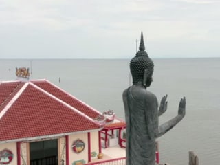Statue hält Hände in die Höhe. Im Hintergrund sieht man ein Gebäude der Tempelanlage. 