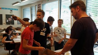 Schüler und Lehrer stehen um eine Kartonkiste, die auf einem Tisch steht und nehmen ihre Handys in Empfang. Schüler und Lehrer stehen um eine Kartonkiste, die auf einem Tisch steht und nehmen ihre Handys in Empfang.