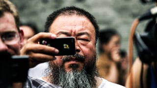 Ai Wei Wei steht in einer Menschenmenge und hält ein Smartphone vor seinem Gesicht.
