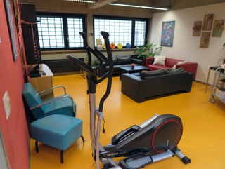 Ein Aufenthaltsraum. Hinten rechts stehen Ledersofas in der Ecke, an der Wand ein TV, im Vordergrund ein Fitnessgerät.