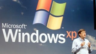 Der ehemalige Microsoft-Chef Bill Gates steht gestikulierend vor einem grossen Windows-XP-Logo.