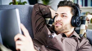 Ein junger Mann mit Dreitagebart liegt auf einem Sofa, trägt Kopfhörer und schaut in sein iPad.