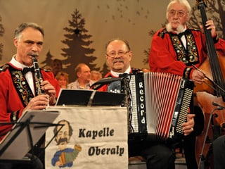 Drei Volksmusikanten in roten Sennenhemden mit Klarinette, Akkordeon und Kontrabass.