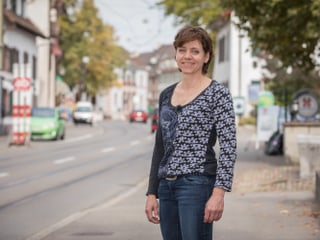 Eien Frau steht auf einer befahrenen Strasse in Riehen und lächelt in die Kamera.