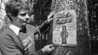 Ein Mann steht neben einem Plakat, dass an einem Baum angebracht ist.