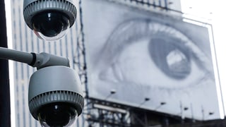 Zwei Überwachungskameras, im Hintergrund ein grosses Plakat, auf dem ein grosses Auge abgebildet ist.
