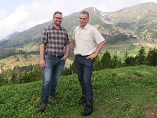 Zwei Männer stehen auf einem Hügel - im Hintergrund Berge und Wälder. 