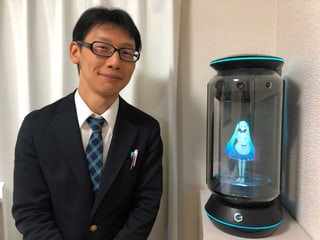 Mann mit Brille neben der Gate-Box, einem Glaszylinder, in dem Miku zu sehen ist. Mann mit Brille neben der Gate-Box, einem Glaszylinder, in dem Miku zu sehen ist.
