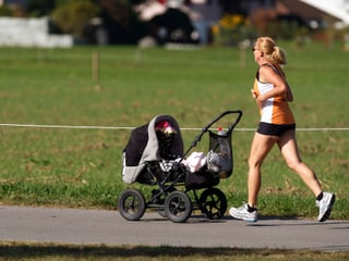 Eine Frau joggt bei strahlender Herbstsonne, mit dem Kinderwagen vor sich, durch den Park.