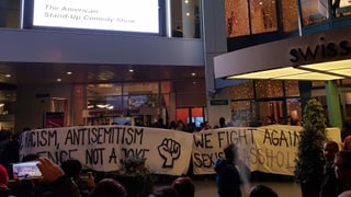 Menschenmenge vor einem Gebäude, einige halten Plakate. Aufschrift: "Racism, Antisemitism... not a joke."