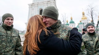 Frau umarmt jungen Mann in militärischer Tarnuniform, im Hintergrund sichtbar ist Kathedrale.