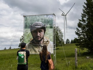 Zwei Menschen stehen im Vordergrund und schauen auf ein Kunstwerk, im Hintergrund steht ein Windrad.