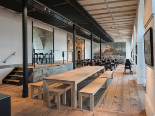 Saal mit langem Holztisch, dahinter kleinere Tische, an einer Wand sieht man einen Rundbogen aus alten Steinblöcken 
