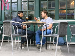 An einem Tisch vor einer Bar sitzen zwei Männer und unterhalten sich. 