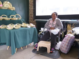 Beverly ist eine ältere schwarze Frau, die auf einem Sessel sitzt. Sie trägt ihre Haare nach hinten gekämmt und einen grauen Pulli mit pinker Schleife.