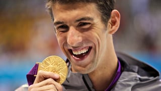 Michael Phelps lächelt und streckt eine Goldmedaille in die Kamera.