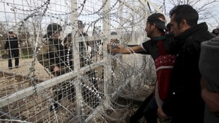Flüchtlinge blicken durch einen Zaun hindurch zwei Polizisten an. 