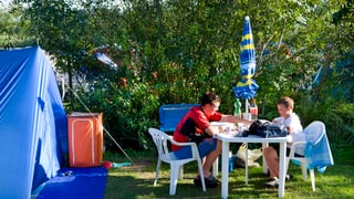 Ein Mann und ein Kind auf weissen Stühlen und an einem weissen Tisch beim Essen auf dem Campingplatz. Daneben ein blaues Zelt. 