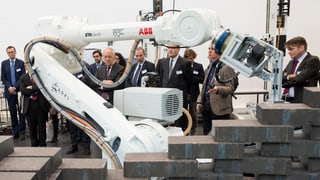 Ein Roboterarm stapelt schwarze Klötze, im Hintergrund schauen Bundesrat Schneider-Ammann und andere Männer interessiert zu.